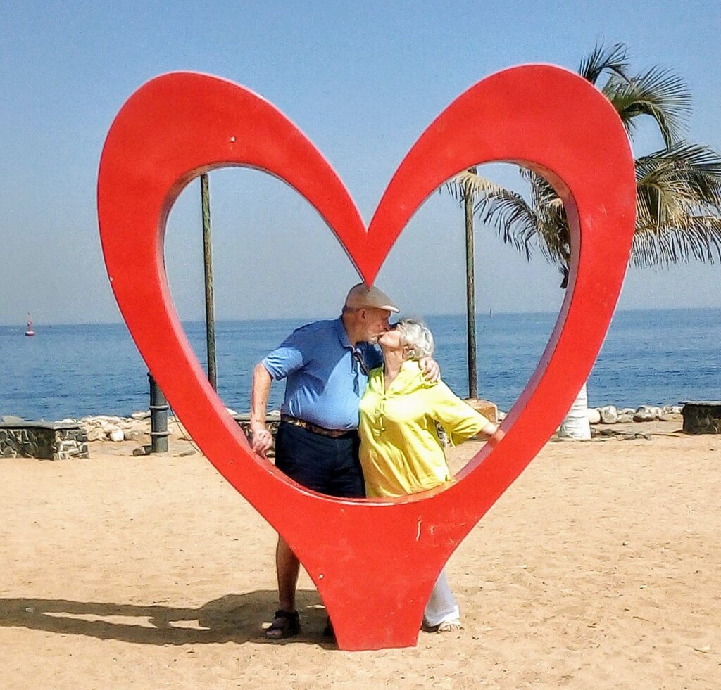 A couple sitting inside a large red heart sculpture by the beach.