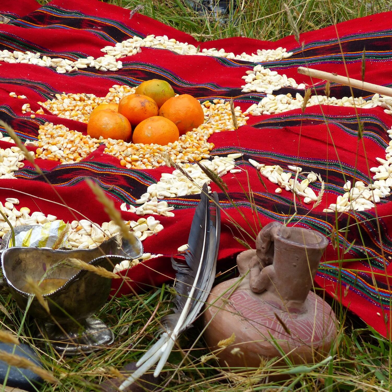 Traditional arrangement with fruits, grains, feathers, and pottery on red cloth.