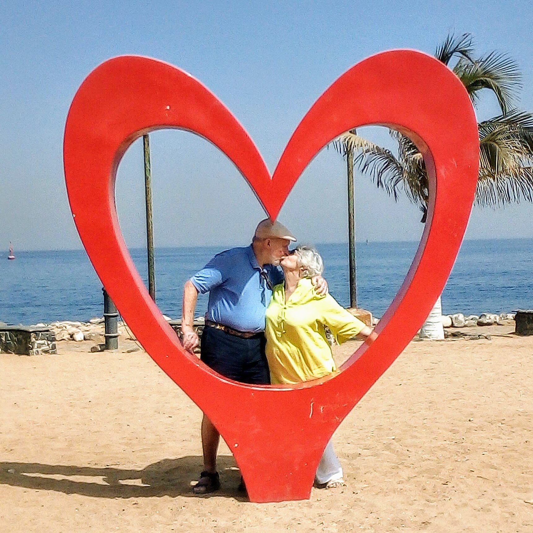 A couple sitting inside a large red heart sculpture by the beach.